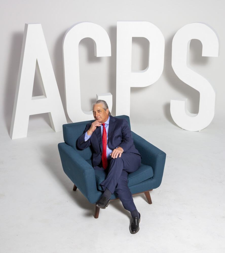 Dr. Patronella sitting in a studio, on an accent chair with the logo ACPS letters behind him